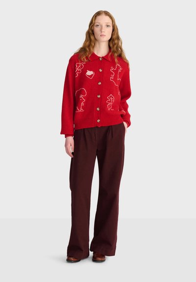 Jeune femme aux longs cheveux roux portant un pull rouge à boutons avec des motifs d’animaux en contour blanc et un pantalon large foncé.
