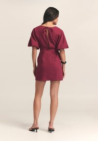Vestido burdeos con mangas cortas abullonadas, detalle de lazo en la espalda, cintura ajustada y longitud por encima de la rodilla. Tejido suave con una textura sutil.