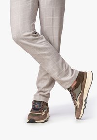 Sneaker in suede marrone e mesh con dettagli crema, suola testurizzata e lacci blu navy, abbinate a pantaloni a quadri chiari.