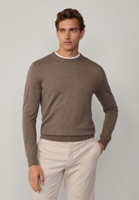 Brązowy sweter dziany z okrągłym dekoltem i długimi rękawami, w połączeniu z jasnymi spodniami. Miękka tekstura i dopasowany krój.