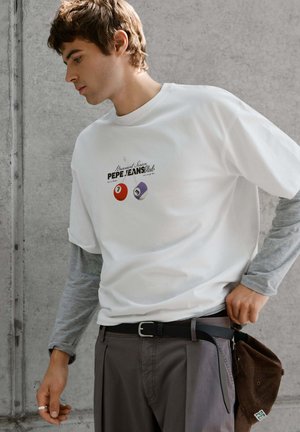 Jeune homme portant un t-shirt blanc Pepe Jeans par-dessus un haut gris à manches longues, ajustant sa ceinture de pantalon, tenant une casquette marron contre un mur gris.