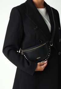 Person klädd i en svart dubbelknäppt rock som håller en svart Mexx axelväska med guldblixtlås och metallnitar mot en vit bakgrund.