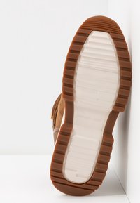 Semelle de basket en suede marron avec une semelle intermédiaire en caoutchouc blanc, présentant un motif texturé et un nom de marque embossé. Le côté a des glands décoratifs.