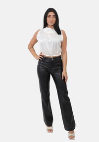 Donna con lunghi capelli scuri che indossa un top senza maniche in satin bianco, pantaloni in pelle nera e sandali bianchi con punta aperta, in piedi con una mano sui fianchi.