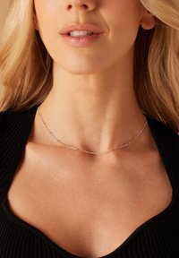 Zilveren ketting met een delicate schakel die kleine ronde en langwerpige schakels bevat, gedragen op blote huid, aangevuld met een zwart geribbeld topje.