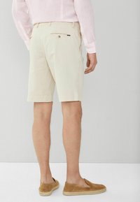 Beige bomuldsshorts har en baglomme med knaplukning. Modellen har brune slip-on sko med en espadrille-lignende sål og opløftede kanter.