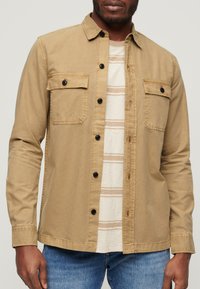 Chemise boutonnée beige avec deux poches poitrine et boutons noirs, portée par-dessus un t-shirt crème rayé, associée à un jean bleu.
