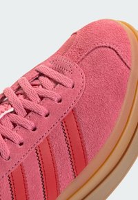 Sapatilha de camurça rosa com atacadores tonais, três riscas vermelhas, acentos texturizados e uma sola de borracha creme com um ligeiro acabamento translúcido.