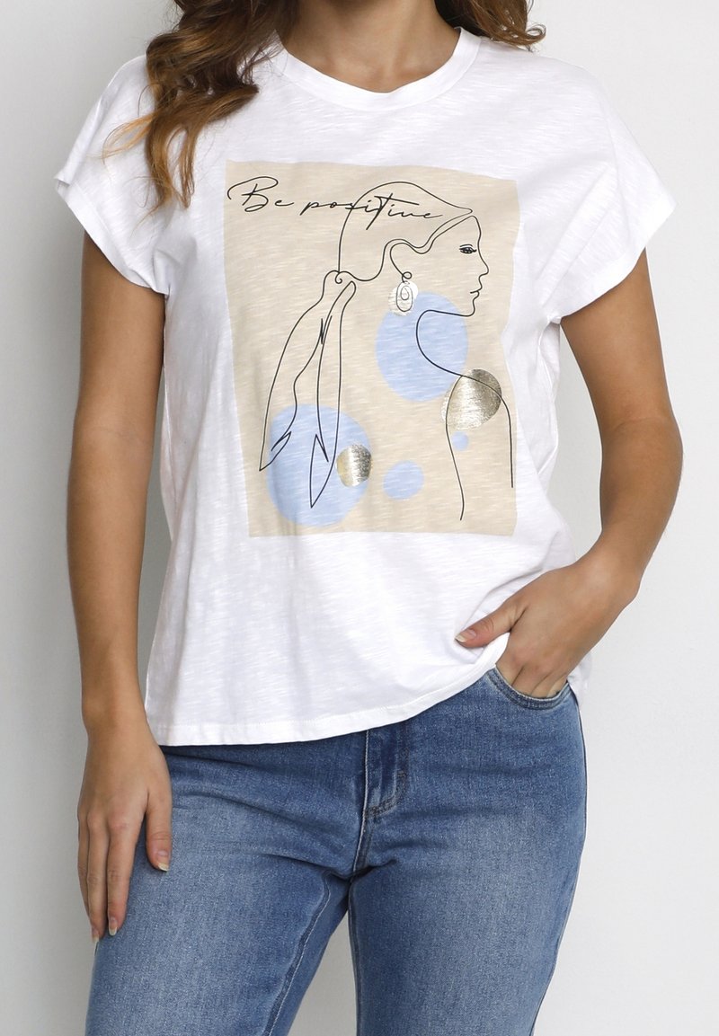 T-shirt blanc avec un dessin linéaire abstrait d'une femme portant des boucles d'oreilles, des cercles bleus, des accents dorés et le texte "Be positive", porté avec un jean bleu.