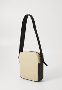 Borsa a tracolla beige e nera dalla forma quadrata, realizzata in materiale sintetico liscio, dotata di tracolla regolabile e chiusura a zip.