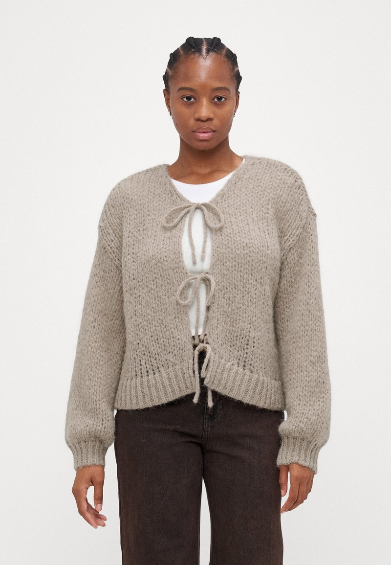 Beige neulecardigan, jossa on löysä istuvuus, sidontakuvio, pitkät hihat ja teksturoitu kangas. Puettuna valkoisen paidan päällä.