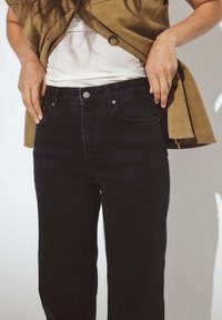 Mustad teksapüksid sirge jalaga, millel on esitaskud ja nupuga sulg. Kantud valge särgi ja beeži välisteki kohal.