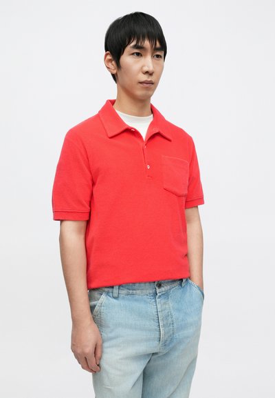 Polo rouge à manches courtes avec col classique et poche poitrine, en tissu texturé. Associé à un jean en denim bleu clair délavé.