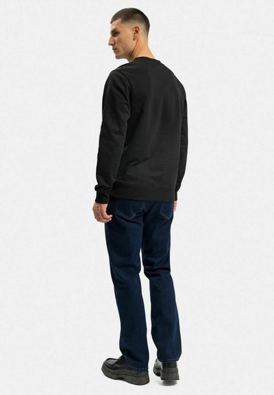 Svart sweatshirt med klassisk rund hals, ribbade ärmslut och midja. Kombinerad med mörkblå jeans och svarta skor. Minimalistisk design.