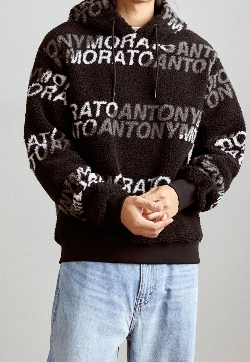 Personne portant un sweat à capuche en polaire noire avec un motif texte blanc et gris « ANTONY MORATO », associé à un jean bleu clair, mains jointes.