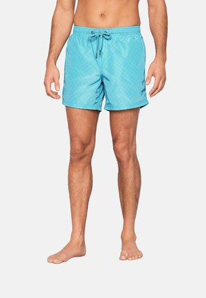 Mann trägt türkisfarbene Badeshorts mit Kordelzug, steht barfuß vor einfachem hellen Hintergrund.