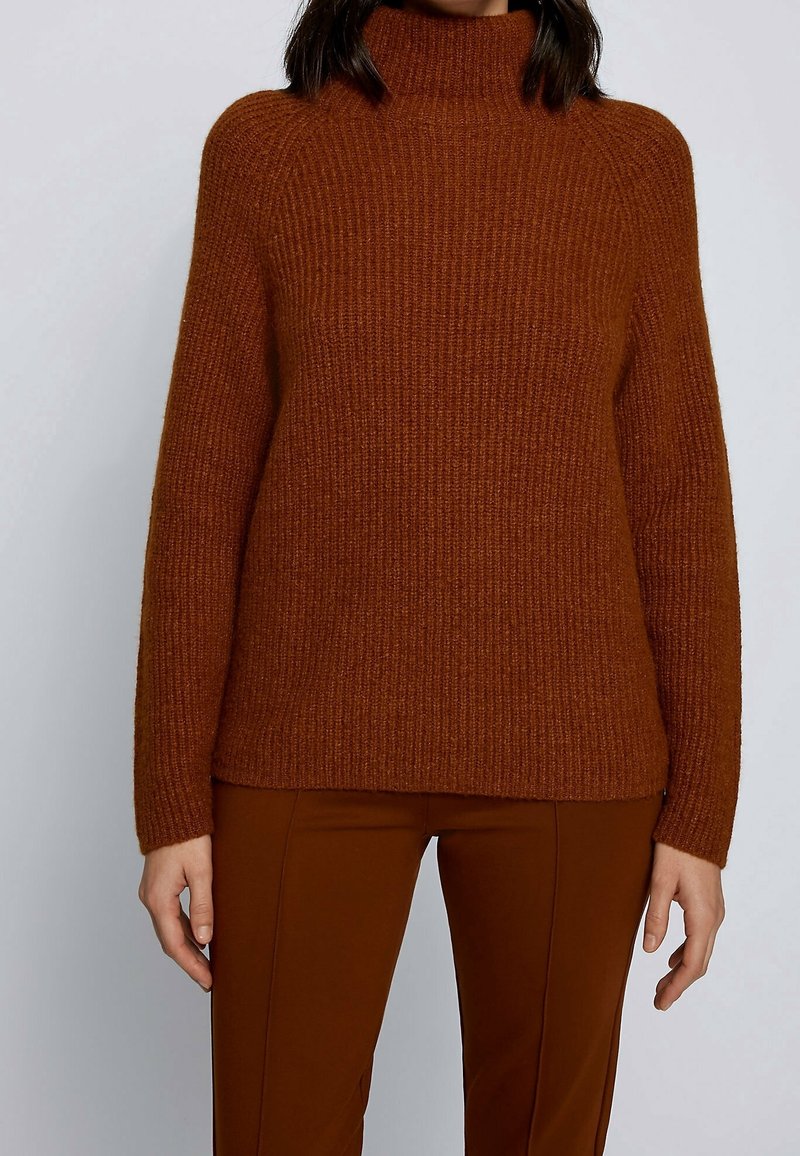 Personne portant un pull à col roulé côtelé marron et un pantalon marron assorti, debout devant un fond clair uni.