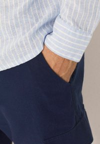 Cămașă în dungi albastre deschise, cu o textură moale, asortată cu pantaloni scurți bleumarin cu un buzunar lateral, punând accent pe confortul și practicitatea îmbrăcămintei casual.