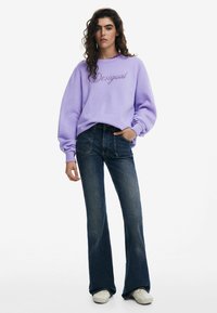 Lila sweatshirt met geborduurd "Desigual" logo, gecombineerd met donkerblauwe flare jeans en witte schoenen. Model heeft krullend haar en houdt handen in de zakken.