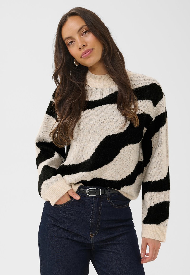 Femme aux longs cheveux bruns porte un pull beige à larges rayures noires et un jean foncé, debout avec une main dans la poche.