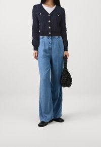 Marineblauer Strickcardigan mit strukturiertem Muster und goldenen Knöpfen, kombiniert mit hellblauen, weit geschnittenen Jeanshosen. Schwarze gewebte Handtasche und flache Schuhe.