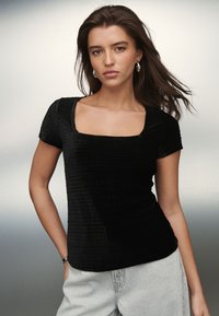 Sort tætsiddende T-shirt med firkantet halsudskæring, korte ærmer og tekstureret stof med ternet mønster, kombineret med lysegrå bukser.