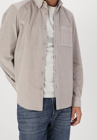Camisa de pana beige claro con botones, un solo bolsillo en el pecho y un corte relajado, llevada encima de una camiseta blanca y combinada con jeans azules.