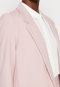 Lichtroze getailleerde blazer met scherpe revers, gladde textuur en subtiele voering. Witte blouse met knopen zichtbaar eronder.