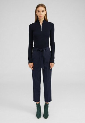 Navy ribbed long-sleeve top with a half-zip collar, paired with high-waisted navy trousers featuring a tied belt. Green ankle boots.