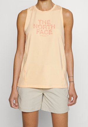 Vrouw die een perzikkleurige mouwloze tanktop van "The North Face" en lichtbeige korte broek draagt, staand tegen een effen achtergrond.