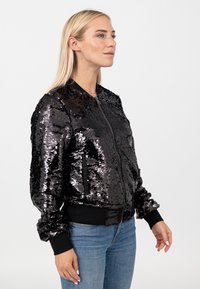 Vrouw met lang blond haar, die een zwarte bomberjas met pailletten en blauwe spijkerbroek draagt, staand tegen een witte achtergrond.