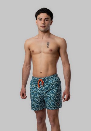 Giovane uomo in piedi senza maglietta che indossa pantaloncini da bagno blu a motivo con coulisse arancione, tatuaggio visibile sul petto sinistro, sfondo grigio neutro.