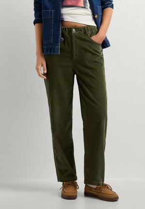 Persoon die olijfgroene corduroy broek, bruine schoenen, een wit crop top en een blauwe spijkerjasje met één hand in de zak draagt.
