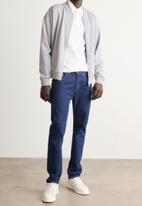 Mand iført en lysegrå zip-jakke, hvid polo t-shirt, blå jeans og hvide sneakers står på et glat gulv foran en ensfarvet væg.