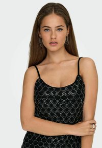 Jeune femme aux longs cheveux bruns, portant un top caraco noir à paillettes et des boucles d'oreilles argentées, posant avec un bras croisé sur sa taille.