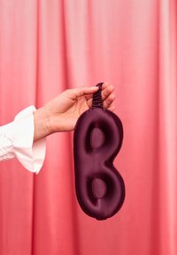 Ręka trzyma fioletową satynową opaskę na oczy na elastycznym pasku na tle różowego gradientu.