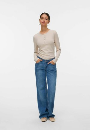 Beige långärmad topp med knäppt halsringning, tillsammans med vida blå jeans. Modellen står med händerna på höfterna och har på sig ljusa skor.