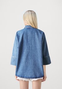 Jeansjacke in Hellblau mit Kragen, Dreiviertelärmel und fransigem unteren Saum. Der Stoff hat eine klassische Textur.