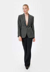 Blazer grigio con texture, colletto a punta, chiusura a un bottone, spalle strutturate e vestibilità su misura, abbinato a pantaloni neri sartoriali.