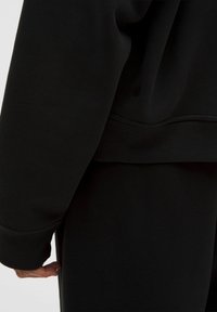 Schwarzer Sweatshirt mit einem strukturierten Stoff, geripptem Saum, leicht übergroßer Passform und einem kleinen Logo-Detail nahe der Naht.