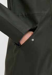 Ciemnozielona wodoodporna kurtka o gładkiej teksturze, z przednią kieszenią zabezpieczoną srebrzystym zatrzaskiem oraz eleganckim, prostym wzorem.