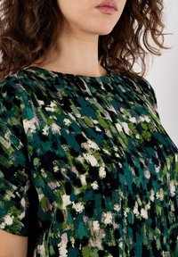 Kurzes Kleid mit einem abstrakten Muster in Grün, Schwarz und Beige. Der Stoff wirkt weich mit einer glatten Textur und hat einen runden Ausschnitt.