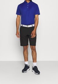 Polo bleu à manches courtes avec trois boutons, associé à un short noir. La tenue est complétée par une ceinture blanche avec boucle argentée et des baskets bleu marine.