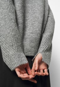 Personne portant un pull gris tricoté ample avec des manches longues couvrant partiellement les mains, qui sont détendues et tenues derrière le dos.