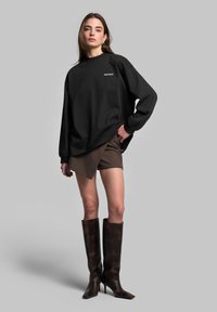Černá oversize mikina, hnědé asymetrické šortky a hnědé vysoké boty do kolen. Hladké textury, minimalistický design a přiléhavé tvary.