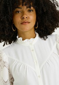 Blouse blanche avec col volanté, boutons à l'avant et tissu texturé. Présente des accents en dentelle sur les manches bouffantes. Boucles d'oreilles en or en forme de cercle.