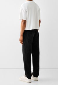 Camiseta blanca de algodón con mangas cortas, combinada con pantalones vaqueros negros de cintura alta y zapatillas de deporte de color claro, visible desde la parte de atrás.