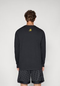 Man with curly hair facing away wearing a black long-sleeve shirt with a small yellow "L" logo on the upper back and patterned black shorts.