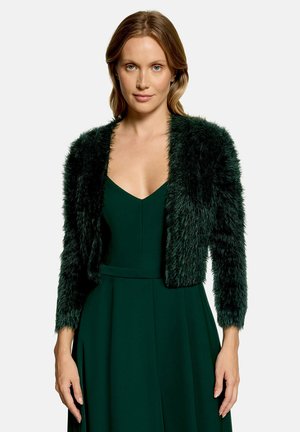 Frau mit hellbraunen Haaren, die ein dunkelgrünes ärmelloses Kleid und eine passende flauschige kurze Strickjacke trägt und nach vorne schaut.
