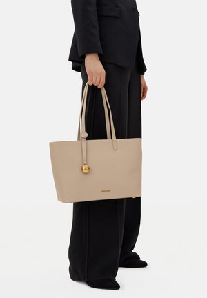 Osoba v černém obleku držící béžovou tašku přes rameno s dlouhými popruhy a zlatým kulatým přívěskem na jednobarevném pozadí.